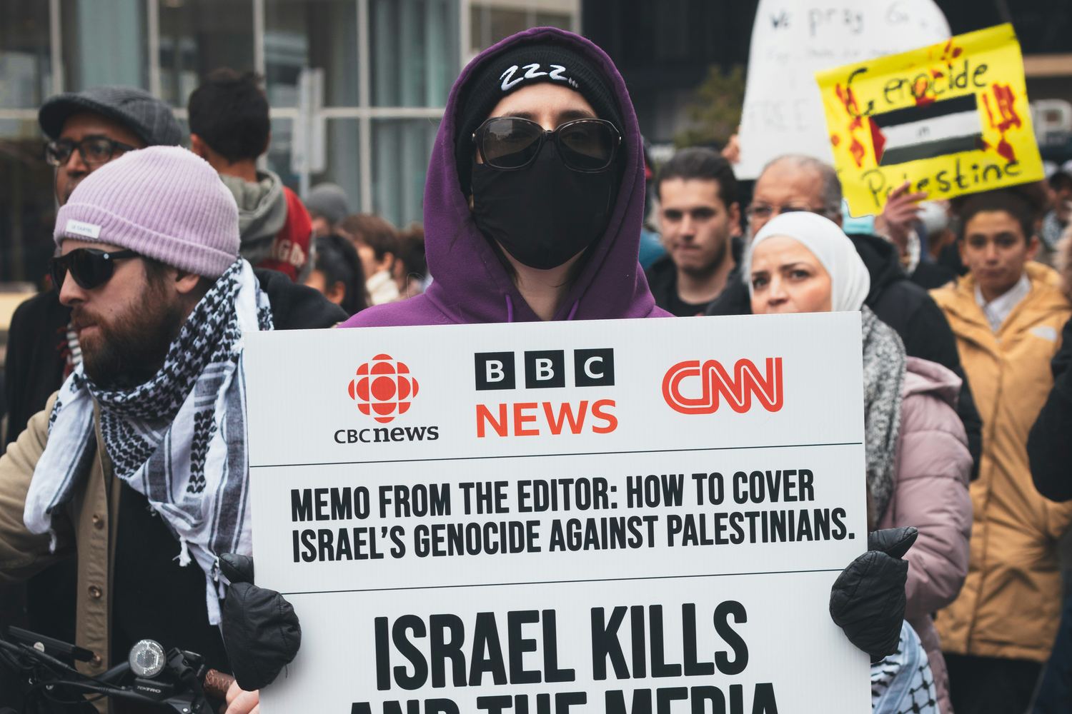 A masked, hooded protester at an anti-Israel demonstration holds a placard accusing Israel of genocide.